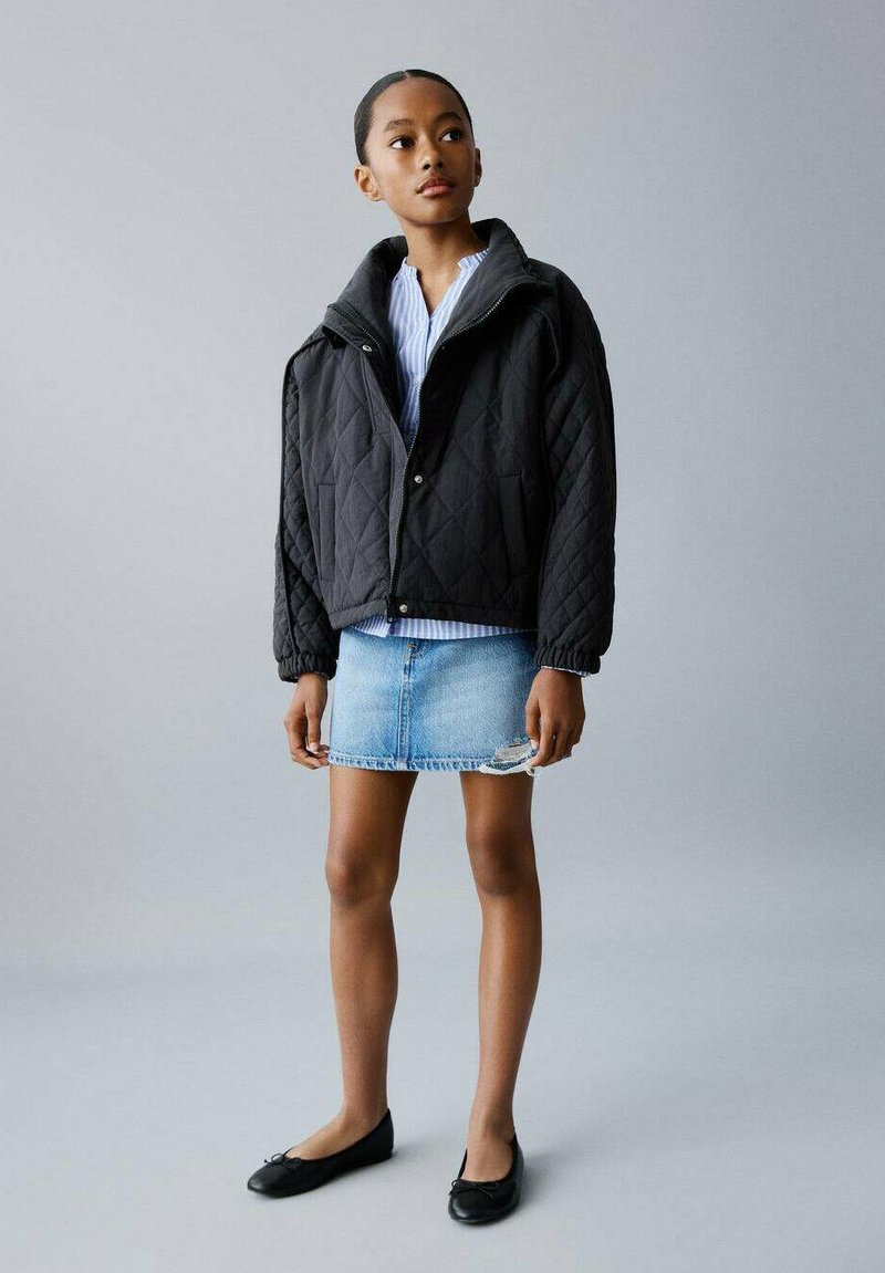 Veste matelassée noire avec un col montant, superposée à une chemise à rayures bleues, associée à une jupe en denim clair et des ballerines noires. Design simple.