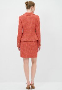 Roter Tweed-Anzug mit taillierter Blazerjacke und Bleistiftrock, der eine Peplum-Taille und goldene Knopfdetails an den Ärmeln aufweist. Glatte Textur.