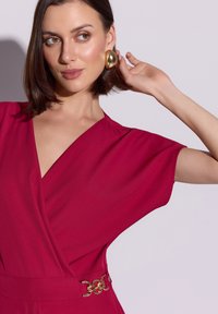 Sukienka kopertowa w kolorze fuksji z krótkimi rękawami, ozdobiona złotym, okrągłym paskiem. Gładki materiał, głęboki dekolt w serek i czyste linie.