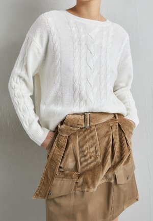 Personne portant un pull en maille torsadée blanc rentré dans une jupe en velours côtelé beige avec une ceinture et des poches avant, mains dans les poches.