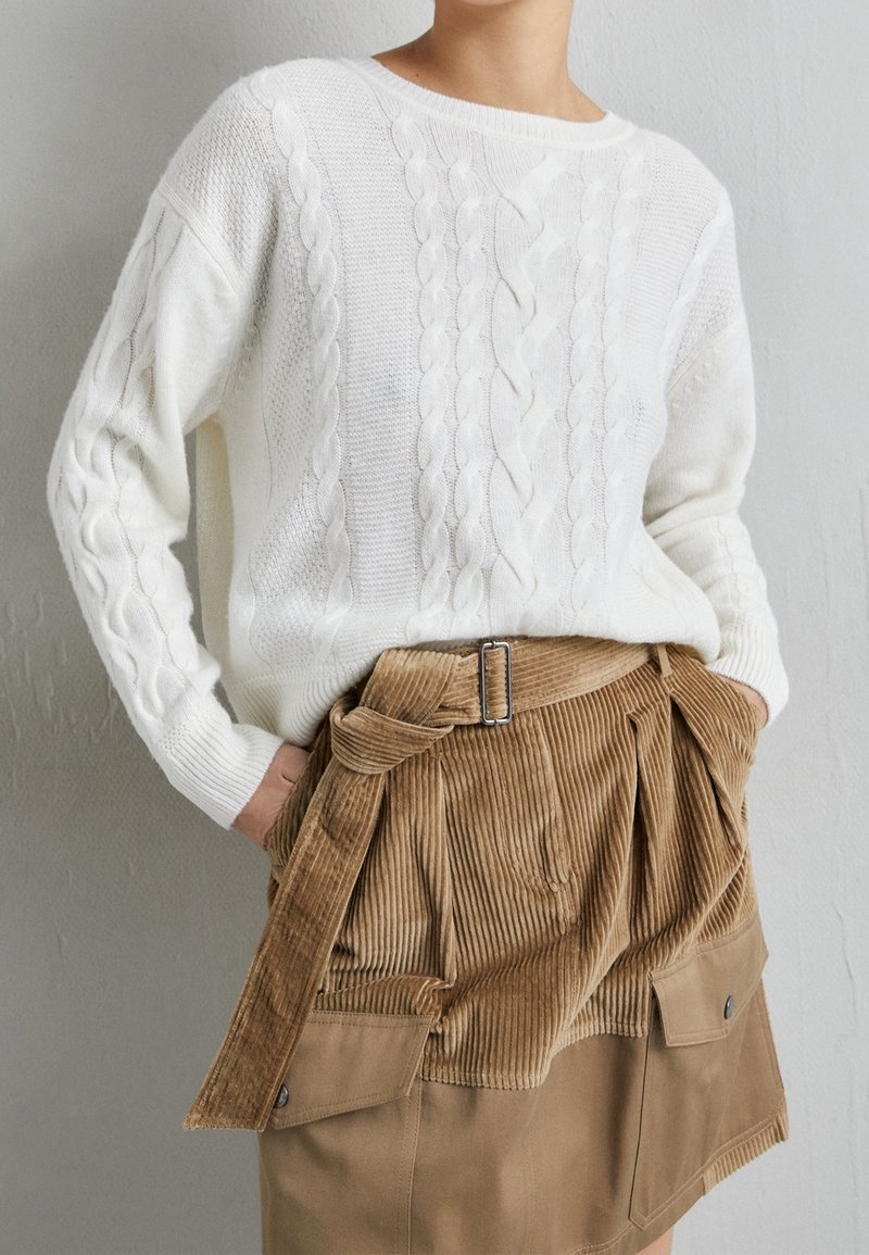 Personne portant un pull en maille torsadée blanc rentré dans une jupe en velours côtelé beige avec une ceinture et des poches avant, mains dans les poches.