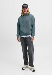 Grauer Kapuzenpullover mit Kordelzug, einer Fronttasche, über einem weißen Shirt getragen. Kombiniert mit dunkelgrauen Jeans und beigen Sneakers.