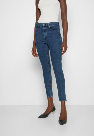 Jeans skinny azul-escuros com cintura alta, apresentando cinco bolsos e fecho de botão, combinados com sapatos de salto agudo pretos.
