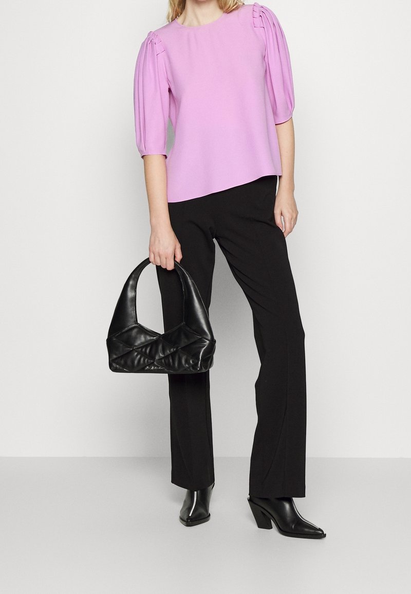 Femme portant un chemisier lavande à manches bouffantes et un pantalon noir, tenant un sac à main en cuir noir, debout devant un fond clair uni.