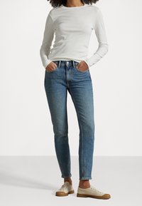 Tajta blå denimjeans med hög midja, kombinerat med en enkel vit långärmad skjorta och beiga snörskor.