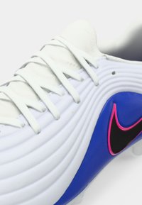 Λευκό ποδοσφαιρικό παπούτσι με υφασμένο επάνω μέρος, μπλε πλευρικές λεπτομέρειες και λογότυπο Nike σε ροζ-μαύρο. Έξι λευκές τρύπες για κορδόνια· στρογγυλεμένη σχεδίαση μύτης.