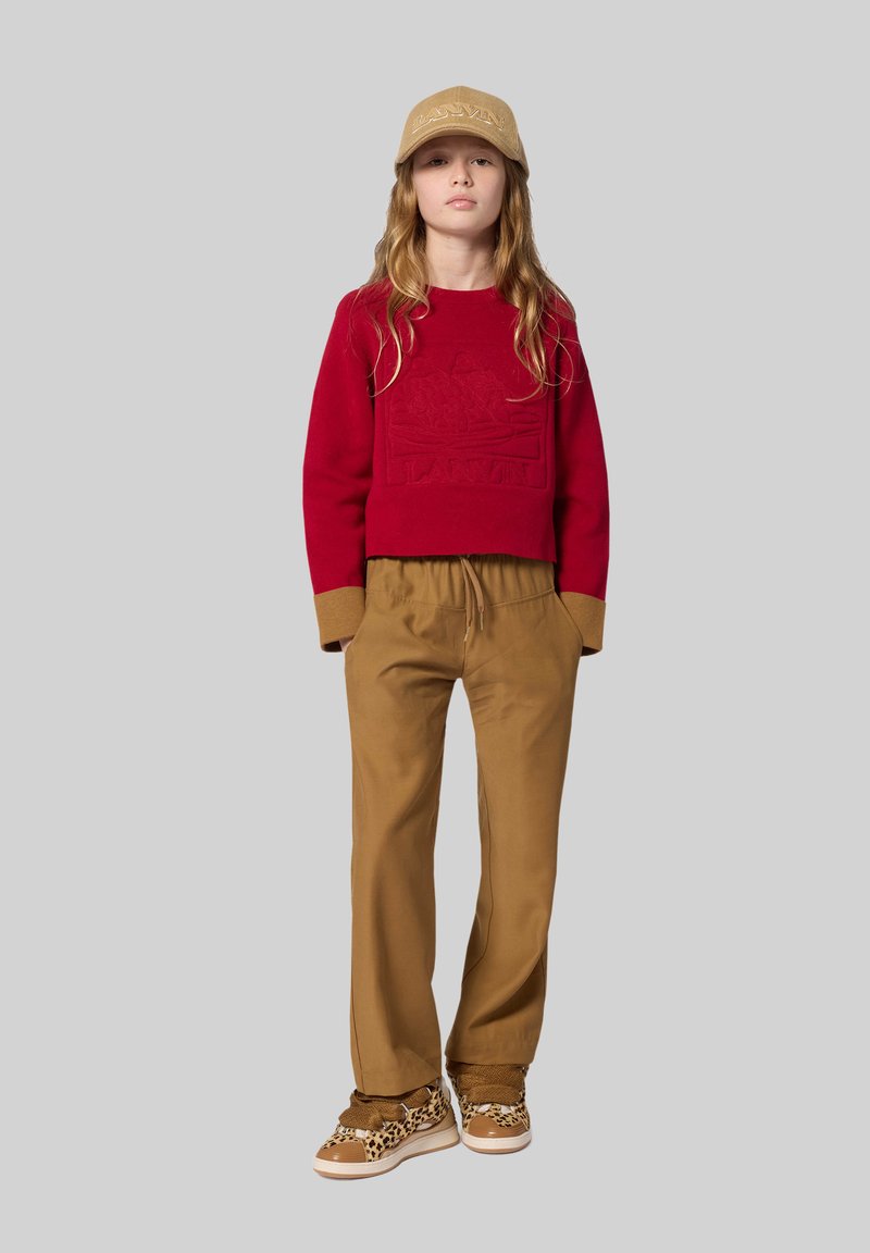 Rode cropped sweatshirt met reliëf logo, beige manchetten, tan trekkoordbroek en sneakers met luipaardprint. Eenvoudige grijze achtergrond.