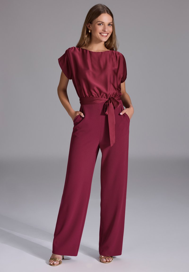 Mauve kombinezon o satynowym wykończeniu, z krótkimi rękawami, ściągniętą talią i szerokimi nogawkami; posiada boczne kieszenie oraz detal z przewiązanym paskiem.