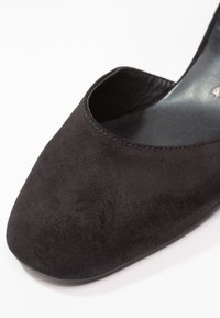 Gros plan sur une chaussure en daim noire avec un bout carré et une texture lisse sur un fond blanc.