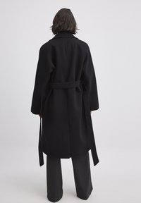 Abrigo negro de lana con cuello, cintura ajustada y mangas largas. Presenta un diseño aerodinámico y un ajuste relajado, que se extiende por debajo de las caderas.