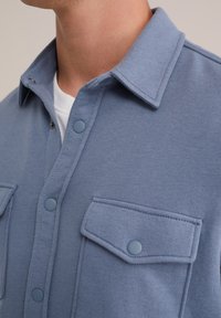 Camisa de forro polar azul con cierre de botones en la parte delantera, dos bolsillos en el pecho y cuello con solapa, que presenta botones a presión azul mate y una textura suave.