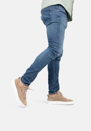 Baskets en daim beige avec des lacets gris clair et une semelle en caoutchouc blanche, associées à un jean en denim bleu. Le design présente un bout arrondi.