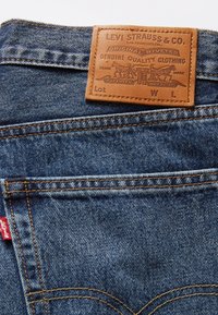 Tmavě modré džíny z denimu s obdélníkovou hnědou koženou nášivkou s vyraženým textem, žlutým prošíváním a malou červenou nálepkou vzadu.