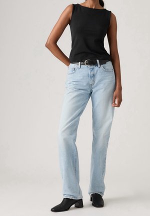 Femme portant un haut noir sans manches, un jean bleu clair droit avec ceinture noire, et des bottines noires à talons, debout avec une main dans la poche.
