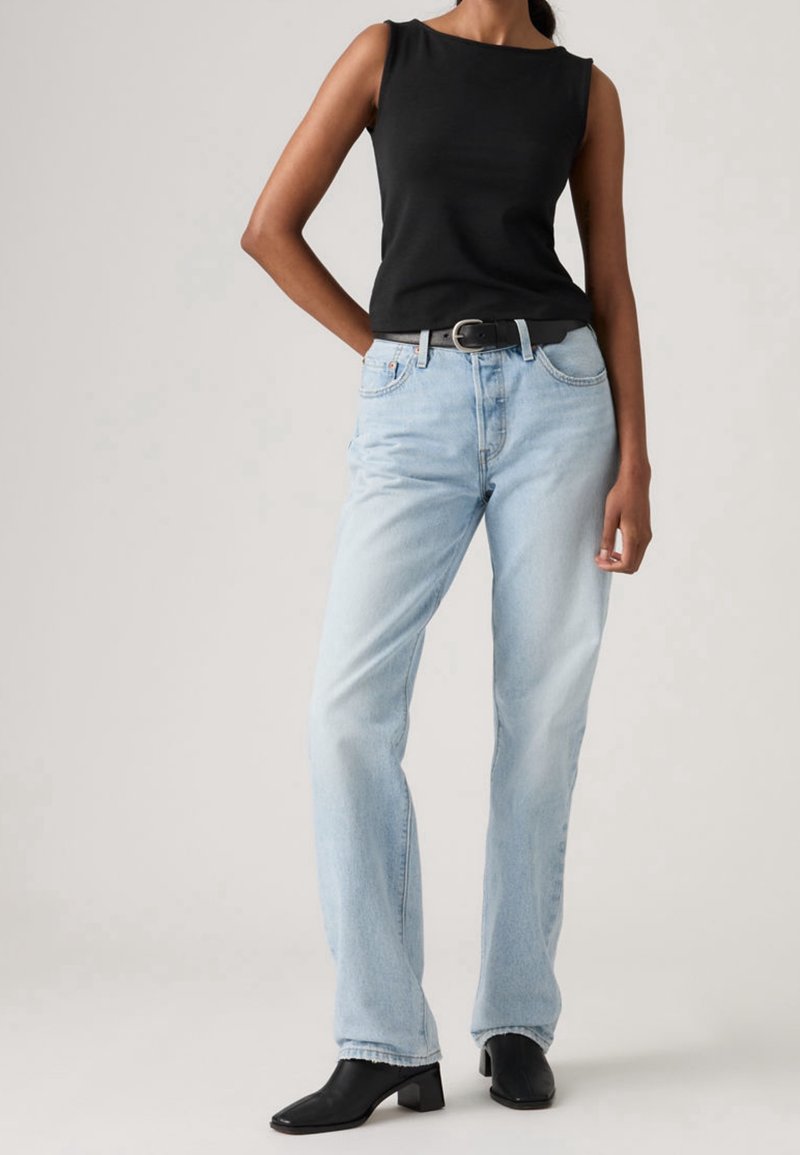 Femme portant un haut noir sans manches, un jean bleu clair droit avec ceinture noire, et des bottines noires à talons, debout avec une main dans la poche.