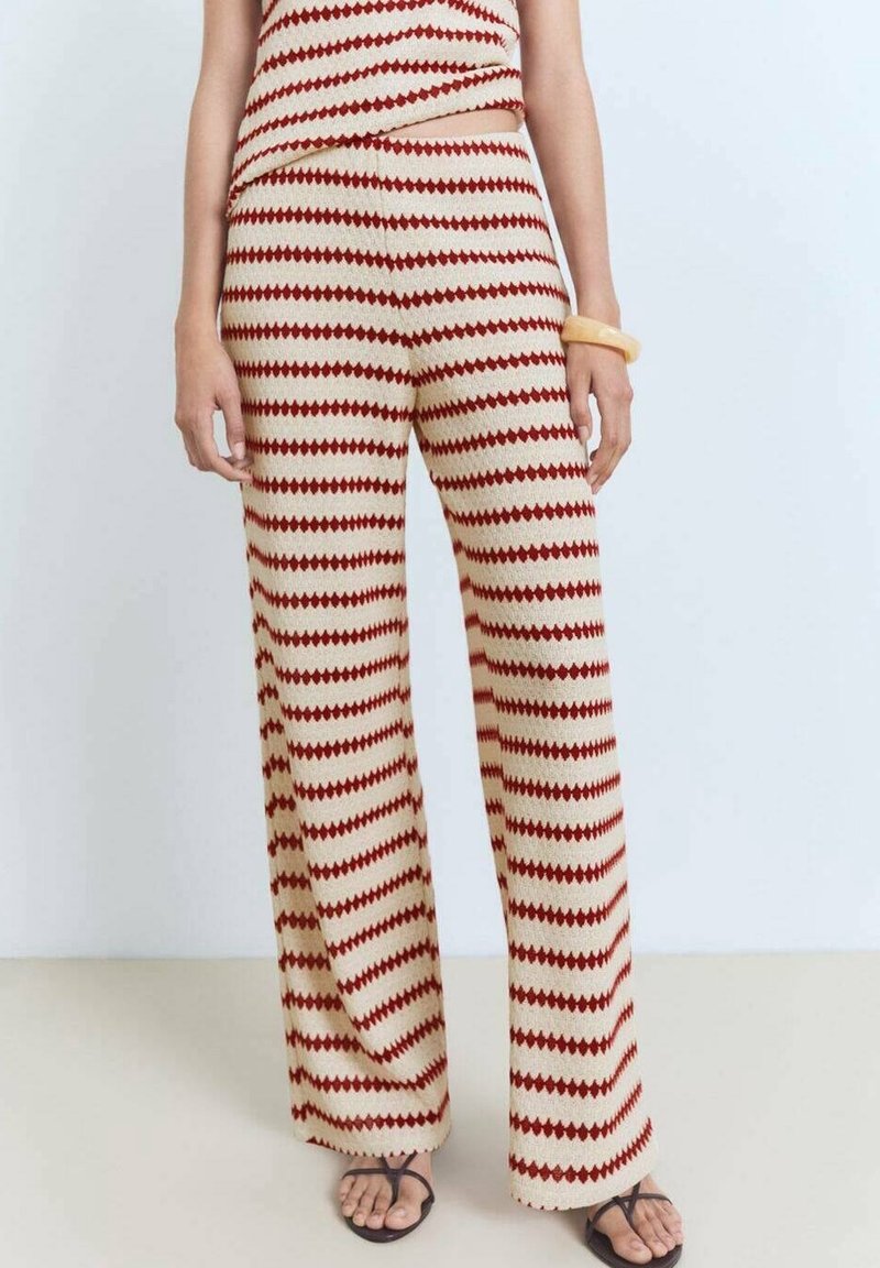 Personne portant un pantalon large en maille beige avec un motif losange rouge, un haut sans manches assorti, des sandales marron et un bracelet beige au poignet droit.