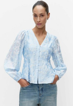 Femme portant un chemisier bleu clair brodé de fleurs avec des manches longues bouffantes et un jean bleu, debout les mains dans les poches.