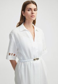 Witte linnen jurk met korte mouwen, voorzien van uitsnijdingen, een V-hals en een gathered taille met een zilverkleurige ronde accent.