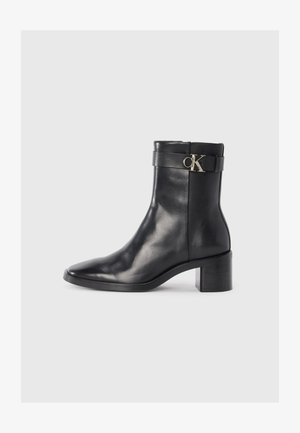 Botte chevillère en cuir noir avec un talon carré, bout pointu et une sangle arborant un logo doré 'CK' sur le côté. Texture et finition lisses.
