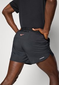 Schwarze Sportshorts mit einem reflektierenden Streifen, Nike-Logo in Koralle und einem strukturierten Bund mit einer Reißverschlusstasche auf der Rückseite.