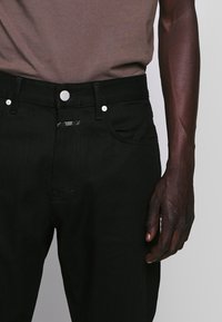 Jeans de denim negro con una silueta ajustada, que presentan un botón metálico, dos bolsillos frontales y sutilezas en el detalle de costura.