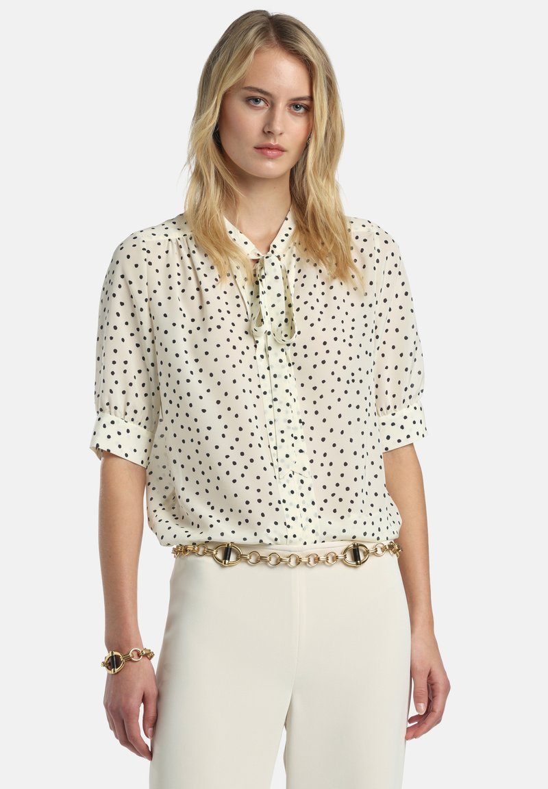 Krém színű blúz fekete pöttyökkel, rövid ujjú, gallérján masni díszítéssel. Világos színű nadrággal és arany láncos övvel párosítva.