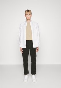 Chemise blanche rayée à boutons au-dessus d'un t-shirt beige, associée à un pantalon en velours côtelé noir et des baskets blanches. Design simple et décontracté.