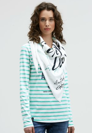 Woman with curly brown hair wearing a white and mint striped long-sleeve shirt and a white scarf with black and turquoise text.
