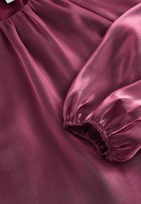 Lila Satinbluse mit glatter Textur, gerafftenÄrmeln und sanftem Glanz, mit entspannter Passform und subtiler Drapierung.