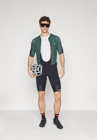 Strój rowerowy składający się z zielonej koszulki z krótkim rękawem, czarnych spodenek z wkładkami z akcentami oraz szarego kasku. Okulary przeciwsłoneczne dopełniają całość.