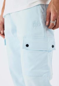 Pantaloni cargo azzurro chiaro con tasche a patta chiuse da bottoni neri, indossati da una persona con un anello a maglie in argento sulla mano destra.