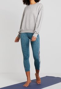 Kvinna står barfota på en yogamatta iklädd en grå sweatshirt och blå beskurna leggings med mönstrade sidpaneler.