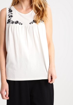 Biały top bez rękawów z czarną kwiatową haftem na górnej części klatki piersiowej, noszony z luźnymi czarnymi spodniami, na jednolitym tle.