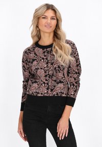 Donna bionda che indossa un maglione a fantasia paisley nero e rosa con polsini e colletto neri, abbinato a jeans neri, in piedi su uno sfondo bianco.
