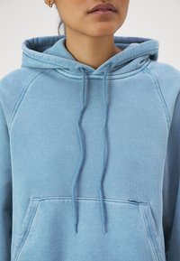 Felpa con cappuccio di un azzurro chiaro, realizzata in un tessuto morbido, dotata di una tasca frontale, lacci nel cappuccio e maniche raglan. Texture liscia, design casual.
