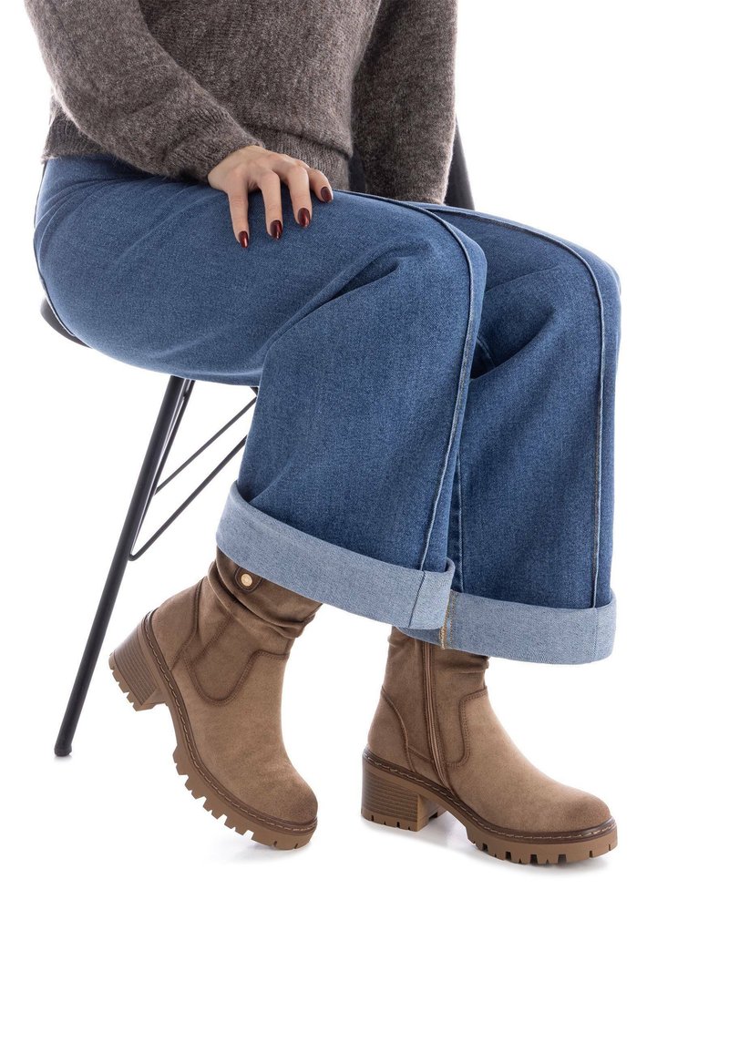 Botas de tobillo de ante marrón con una suela robusta, combinadas con vaqueros de denim azul de pierna ancha con los puños doblados, sentadas en una silla negra.