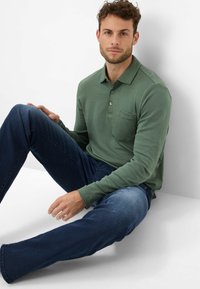 Langärmliges grünes Poloshirt mit einer Brusttasche, kombiniert mit dunkelblauen Jeans, sitzend vor einem weißen Hintergrund.