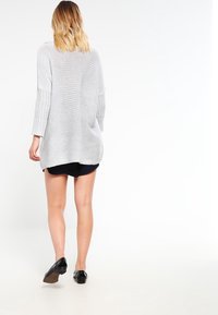 Jasnoszary, oversize'owy sweter z dzianiny z fakturowanymi, prążkowanymi rękawami, otwartym plecami i luźnym krojem, zestawiony z ciemnymi szortami i czarnymi butami.