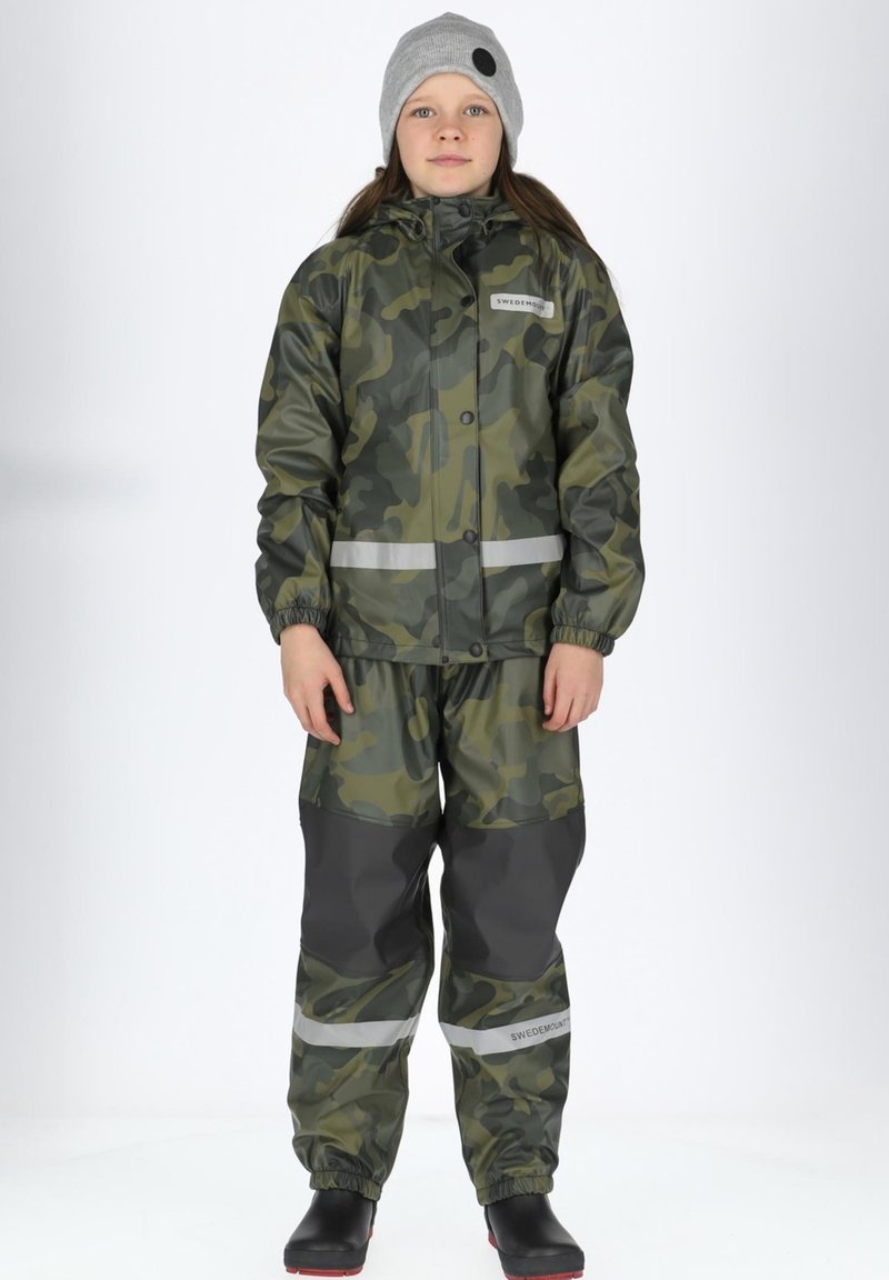 Grøn camo regnjakke og bukser med reflekterende striber. Elastiske manchetter og talje, parret med sorte gummistøvler og en grå hue.