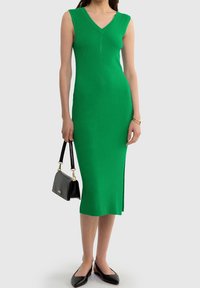Robe tricotée verte côtelée avec un décolleté en V, sans manches, longueur midi, fente sur le côté, associée à un sac à main noir et des chaussures plates.