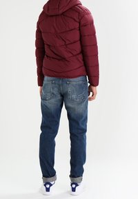 Burgunderfärgad puffjacka med kviltat mönster, jeans i blått med urblekta lår, synliga fickor och vita sneakers med blå accenter.
