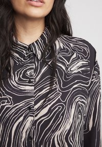 Blusa nera e crema con un motivo a linee ondulate, apertura con bottoni sul davanti e colletto classico. Realizzata in un tessuto morbido con una leggera lucentezza.