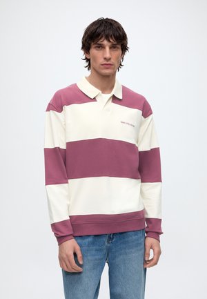 Man wearing long-sleeve rugby shirt with wide horizontal cream and mauve stripes and blue jeans, standing against plain white background.