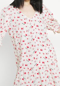 Top floreale con scollo a V, maniche corte a sbuffo e tessuto bianco leggero. Presenta stampe di fiori rosa e rossi con foglie verdi.