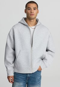 Felpa grigia con zip, dotata di un ampio cappuccio e tasche frontali, realizzata in un morbido tessuto testurizzato. Presenta polsini a coste e una vestibilità rilassata.