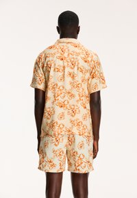 Bloemenoverhemd met korte mouwen en bijpassende shorts van lichtgewicht stof; crèmekleurige achtergrond met een levendig oranje bloemenpatroon.