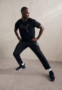Juodas trumpomis rankovėmis marškinėlis su baltu Puma logotipu, derinamas su juodomis sportinėmis kelnėmis ir juodais sportiniais batais su raudonais akcentais bei baltais sokais.