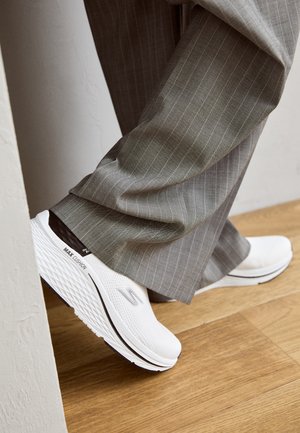 Białe sneakersy z teksturowaną siateczką na cholewce, czarnym akcentem i grubą, wyściełaną podeszwą. W połączeniu z szarymi spodniami w prążek.