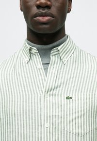 Zöld-fehér csíkos, gombos ing gallérral, mellzsebbel és egy kis hímzett logóval. Puha anyag, függőleges mintával.