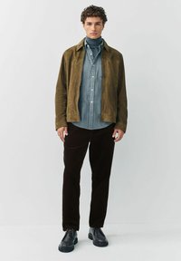 Young man standing wearing a brown suede jacket, blue striped shirt over a blue turtleneck, black pants, and black leather shoes.